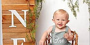 Arlo is registered to the contest to win money with this photo: baby, child, smile, wicker_chair, romper, greenery, vines, wooden_sign, one, birthday, portrait, studio, cute, infant, sitting, happy, decor, white_background, celebration, small_chair