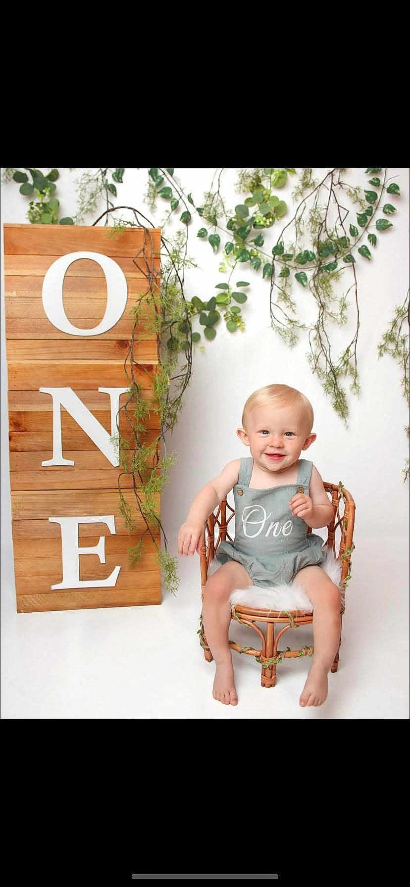 Arlo is registered to the contest to win money with this photo: baby, child, smile, wicker_chair, romper, greenery, vines, wooden_sign, one, birthday, portrait, studio, cute, infant, sitting, happy, decor, white_background, celebration, small_chair
