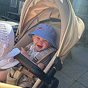 Arlo is registered to the contest to win money with this photo: baby, stroller, hat, smiling, child, sunlight, outdoor, happy, seatbelt, bucket_hat, beige, fabric, toy, sunshade, infant, cute, baby_clothes, seat, baby_boy, laughing
