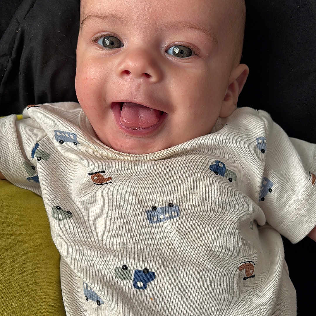 Kailum is registered to the contest to win money with this photo: baby, black_background, child, closeup, clothing, cushion, cute, eyes, face, green_cushion, happy, head, infant, mouth, onesie, portrait, skin, smiling, tongue_out, vehicle_print