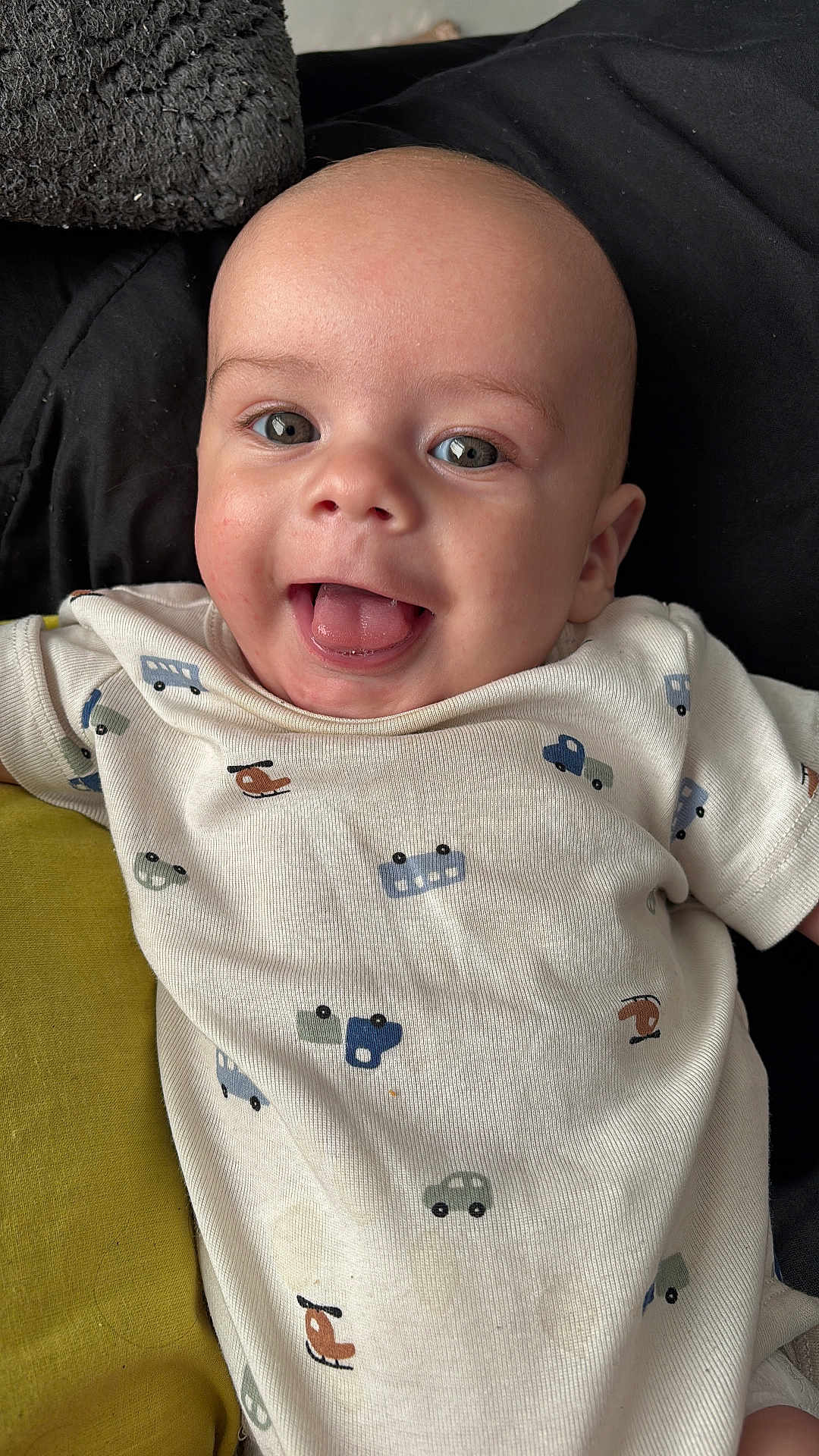Kailum is registered to the contest to win money with this photo: baby, smiling, tongue_out, onesie, vehicle_print, infant, happy, cute, child, face, head, clothing, cushion, black_background, green_cushion, portrait, closeup, skin, eyes, mouth
