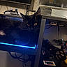 animal, black_cat, cables, cat, close_up, computer_accessories, dark_fur, desk, electronics, feline, gaming_controller, home_office, indoor, led_light, paw, pet, relaxed, shelf, technology, yellow_eyes