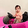 toddler, child, toy_car, pink, steering_wheel, indoor, wall, seat, clothing, person, young_child, play, vehicle, serious, sitting, hair, face, eyes, hands, footwear