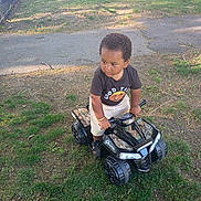 Messiah is registered to the contest to win money with this photo: child, toy_vehicle, grass, tree, fence, parked_car, outdoor, daylight, clothing, shoes, expression, play, vehicle, nature, sidewalk, park, person, young_child, casual, sunlight