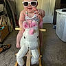 baby, child, toddler, sunglasses, smiling, unicorn, rocking_horse, plush_toy, toy, indoor, bedroom, closet, laundry_basket, carpet, door, clothing, seated, portrait, playtime, funny