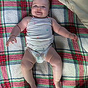 Josie is registered to the contest to win money with this photo: baby, infant, child, smiling, lying_down, portrait, face, eyes, arms, legs, diaper, onesie, blanket, plaid, bedding, pillow, toy, indoor, happy, cozy