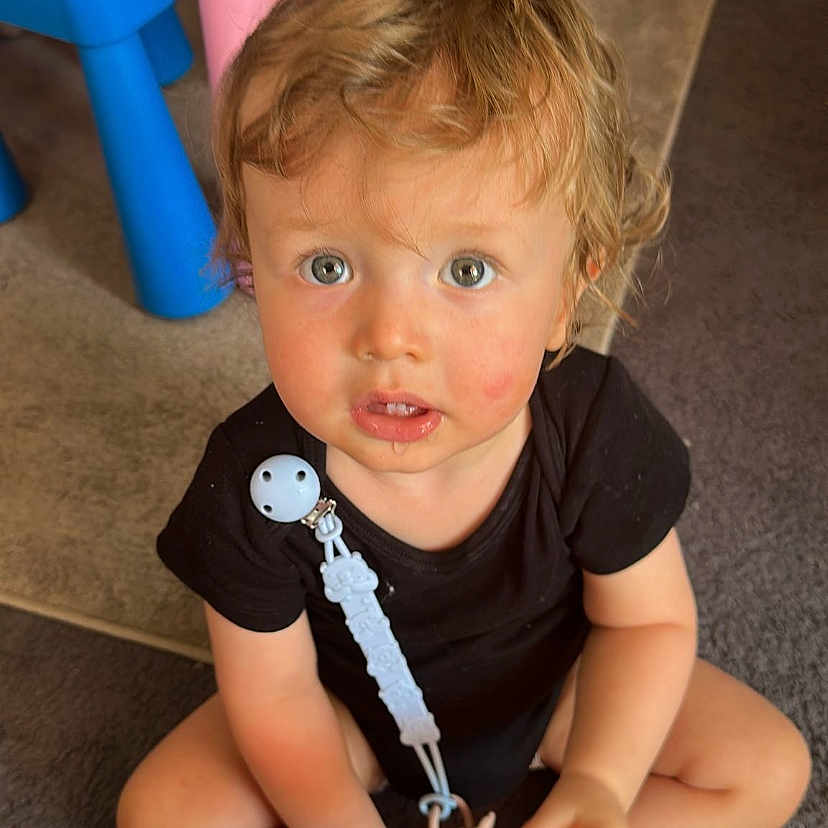 Tyler participe au concours pour gagner de l'argent avec cette photo : baby_accessory, black_clothing, blond_hair, blue, blue_eyes, carpet, child, curious, curly_hair, floor, indoor, pacifier_clip, pink, plastic_chair, playtime, sitting, toddler, toy, toy_bowl, toy_crib