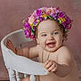 baby, flower_crown, smiling, chair, pink_background, happy, child, portrait, cute, skin, jewelry, gold_chain, earrings, curly_hair, indoor, cute_expression, playful, holding, seated, photography