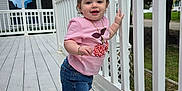 Kimberly is registered to the contest to win money with this photo: toddler, child, standing, pink_shirt, denim_shorts, sneakers, white_railing, wooden_deck, outdoor, house, greenery, cloudy_sky, bracelet, necklace, hair_bows, smiling, person, daylight, casual_clothing, suburban