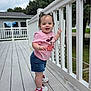 toddler, child, standing, pink_shirt, denim_shorts, sneakers, white_railing, wooden_deck, outdoor, house, greenery, cloudy_sky, bracelet, necklace, hair_bows, smiling, person, daylight, casual_clothing, suburban