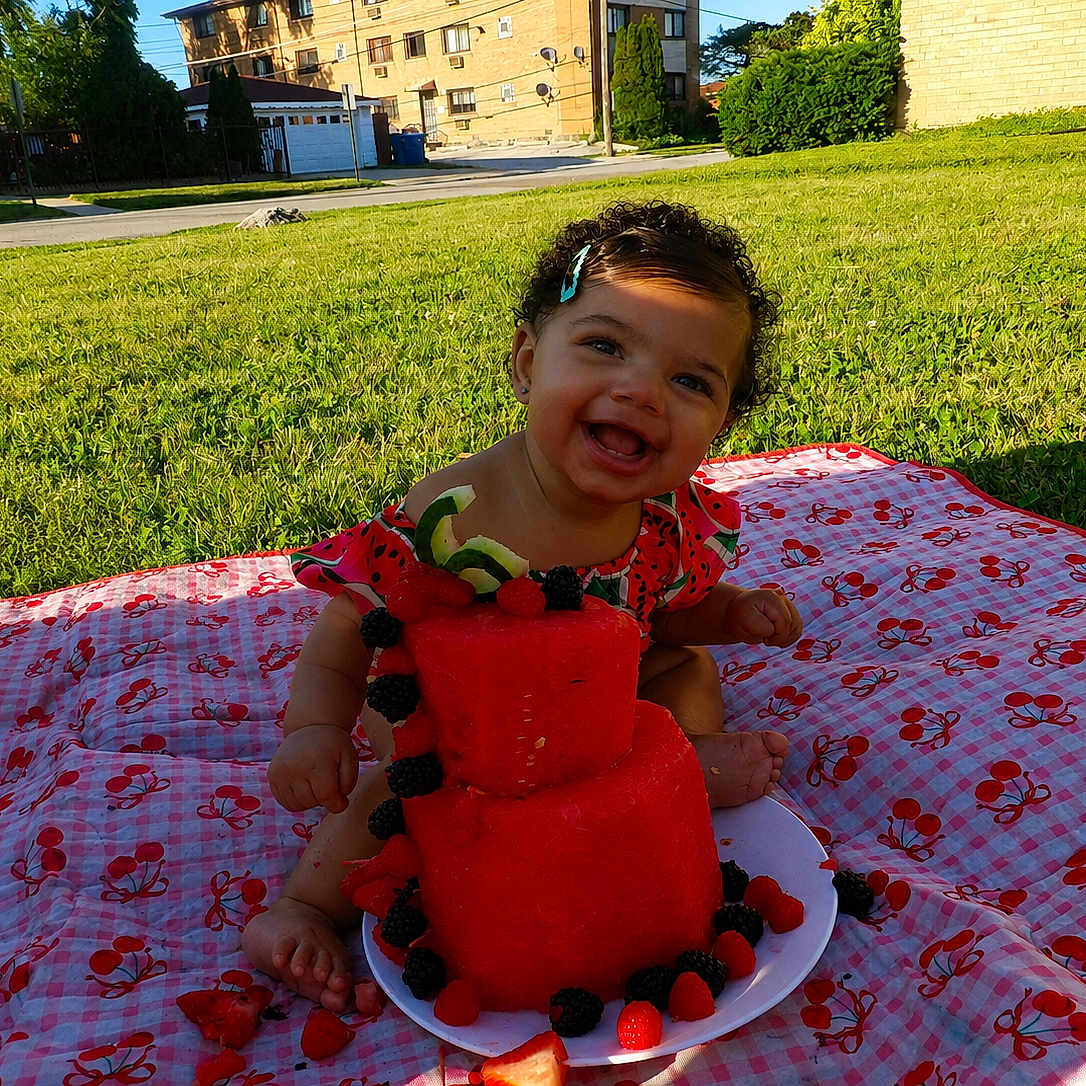 Amira joined the competition — help win amazing prizes! baby, child, smiling, curly_hair, hair_clip, watermelon_cake, berries, picnic_blanket, cherry_pattern, outdoor, grass, sunlight, plate, happy, sitting, summer, food, fruit, nature, portrait