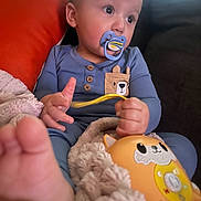 Hudson is registered to the contest to win money with this photo: baby, pacifier, toy, blanket, pajamas, couch, orange_cushion, child, foot, hand, face, indoor, plush, comfort, cute, infant, relaxing, portrait, young_child, blue_clothing
