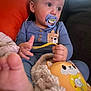 baby, pacifier, toy, blanket, pajamas, couch, orange_cushion, child, foot, hand, face, indoor, plush, comfort, cute, infant, relaxing, portrait, young_child, blue_clothing