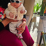 Constance a rejoint le concours — aidez-le/la à gagner de superbes lots ! baby, sunglasses, vest, adult, lap, indoor, couch, plant, sunlight, window, potted_plant, floor, tile_floor, hand, pants, slippers, person, fashion, comfort, relaxation