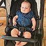 baby, child, stroller, denim_dress, indoor, floor_tiles, baby_boy, seatbelt, black_stroller, baby_clothing, infant, baby_face, cute, child_seated, furniture, wooden_table, footwear, baby_hands, baby_feet, expression