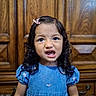 blue_dress, child, clothing, curly_hair, cute, dark_hair, face, front_facing, girl, happy, indoor, looking_at_camera, open_mouth, photography, pink_bow, portrait, smiling, standing, toddler, wooden_background