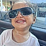 braces, brown_hair, car, car_interior, child, close_up, floral_design, happy, long_hair, outdoor, pink_shirt, playful, portrait, seated, smiling, storefront, sunglasses, sunlight, teeth, window