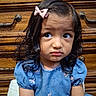 blue_dress, child, close_up, dark_hair, drawer, dresser, embroidered_dress, face, girl, indoors, looking_at_camera, pink_bow, portrait, portrait_photography, serious_expression, sitting, soft_toy, toddler, warm_lighting, wooden_furniture