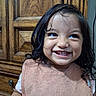 bib, brown_eyes, child, close_up, curly_hair, dark_hair, face, furniture, girl, happy, indoor, joy, long_hair, photography, pink_clothing, portrait, smiling, teeth, toddler, wooden_door