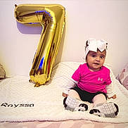 Rayssa Marya a rejoint le concours — aidez-le/la à gagner de superbes lots ! baby, balloon, blanket, celebration, child, cute, decor, floor, gold_balloon, happy, headwear, indoors, number_seven, pink_shirt, portrait, shoes, sitting, toddler, wall, white_bow