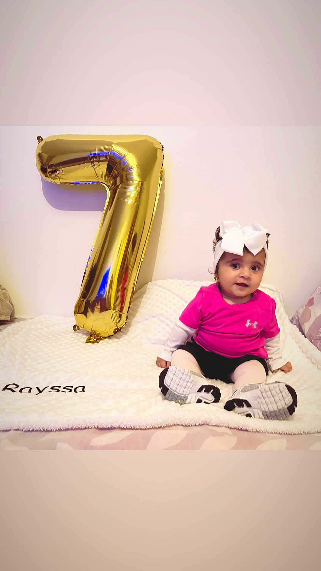 Rayssa Marya a rejoint le concours — aidez-le/la à gagner de superbes lots ! baby, balloon, blanket, celebration, child, cute, decor, floor, gold_balloon, happy, headwear, indoors, number_seven, pink_shirt, portrait, shoes, sitting, toddler, wall, white_bow