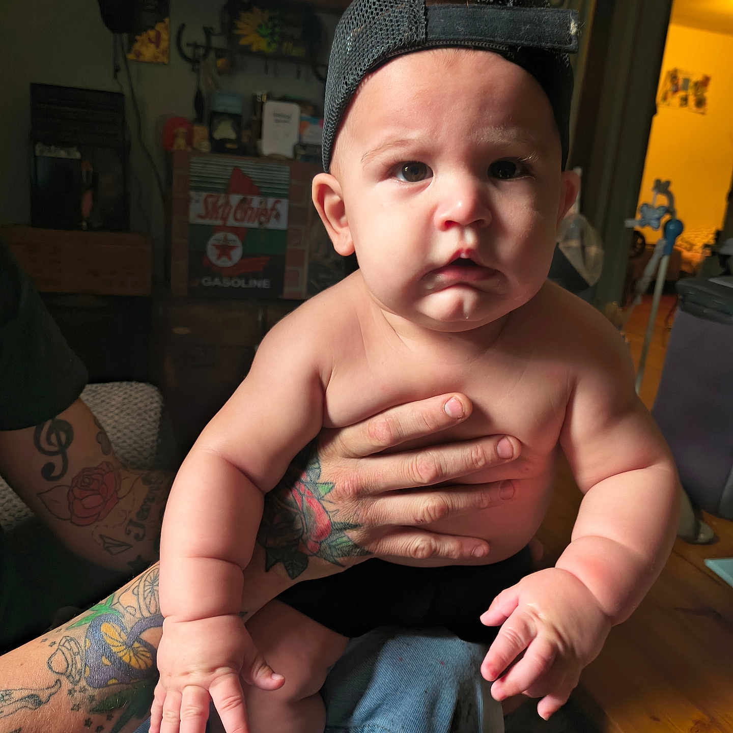 Myles is registered to the contest to win money with this photo: baby, child, tattoo, cap, indoor, person, hand, skin, furniture, clothing, sitting, face, expression, arm, wooden_floor, decor, room, light, curious, holding