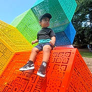 Logan joined the competition — help win amazing prizes! blue, boy, cap, casual_clothing, child, colorful_cubes, daytime, geometric_pattern, grass, green, orange, outdoor, park, playground, sculpture, sitting, sneakers, sunny, trees, yellow