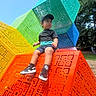 blue, boy, cap, casual_clothing, child, colorful_cubes, daytime, geometric_pattern, grass, green, orange, outdoor, park, playground, sculpture, sitting, sneakers, sunny, trees, yellow
