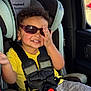 toddler, child, car_seat, sunglasses, yellow_shirt, seatbelt, car_interior, smiling, curly_hair, hand, window, clothing, babytrend, safety_harness, playful, seat, child_safety, casual, fun, portrait