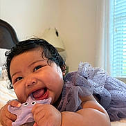 Zipporah is registered to the contest to win money with this photo: baby, child, infant, toy, starfish_toy, purple_dress, curly_hair, smiling, chewing, soft_surface, bedroom, lamp, window, blinds, happy, closeup, portrait, indoor, cute, playful