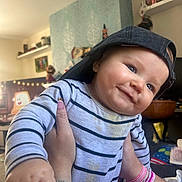Teddy joined the competition — help win amazing prizes! baby, child, smiling, striped_shirt, cap, backwards_cap, hand, bracelet, tattoo, indoor, room, furniture, decorations, person, portrait, happy, face, closeup, infant, playful