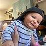baby, child, smiling, striped_shirt, cap, backwards_cap, hand, bracelet, tattoo, indoor, room, furniture, decorations, person, portrait, happy, face, closeup, infant, playful
