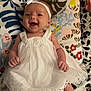 baby, barefoot, blanket, cheerful, child, colorful_background, cute, female, floral_pattern, happy, headband, indoors, infant, lying_down, portrait, small_hands, smiling, soft_lighting, white_dress, young_child