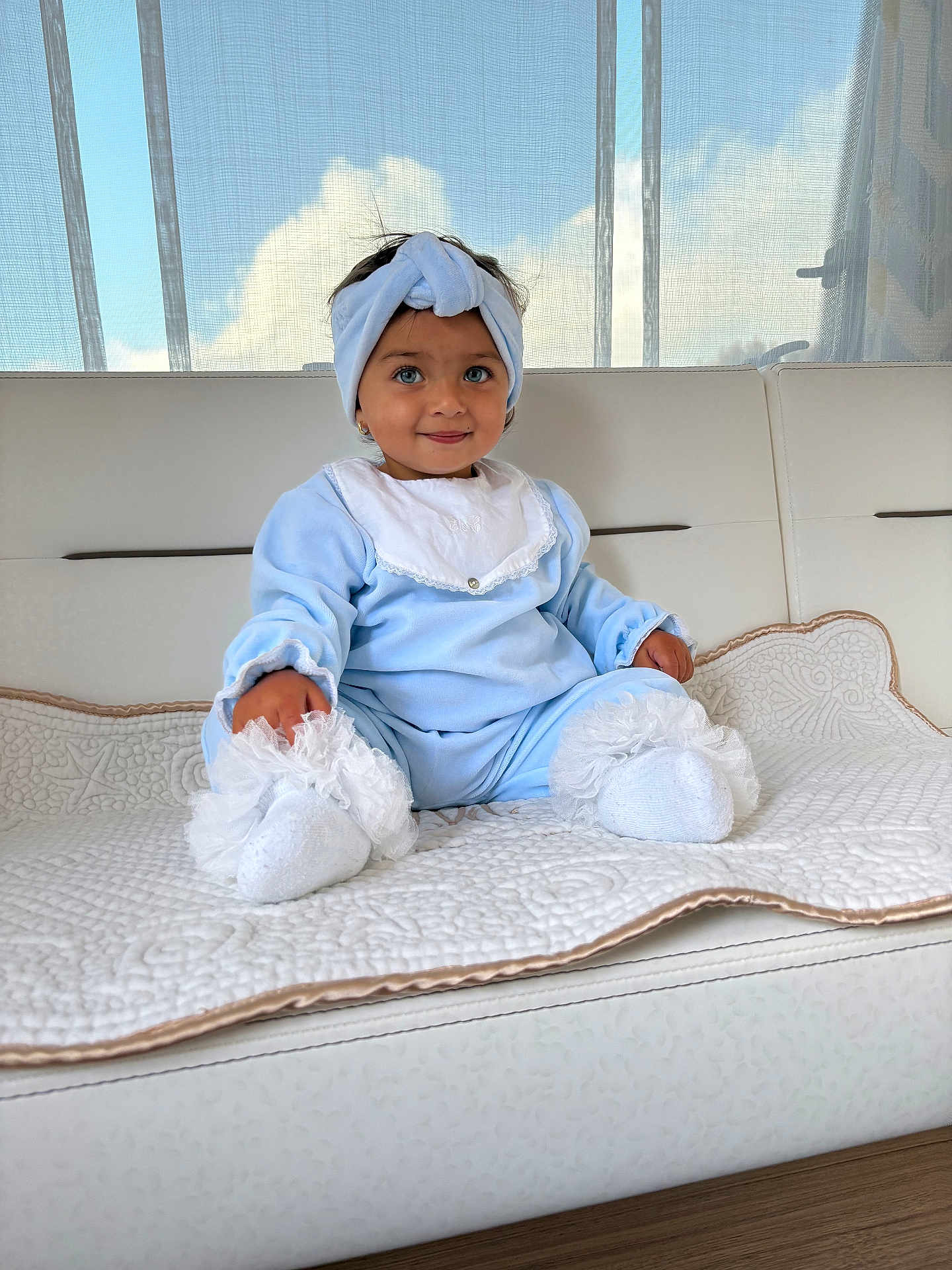 Eva participe au concours pour gagner de l'argent avec cette photo : toddler, child, blue_outfit, headband, white_socks, quilted_blanket, couch, indoor, curtains, window, sky, clouds, smile, cute, portrait, sitting, soft_lighting, baby, homedecor, person