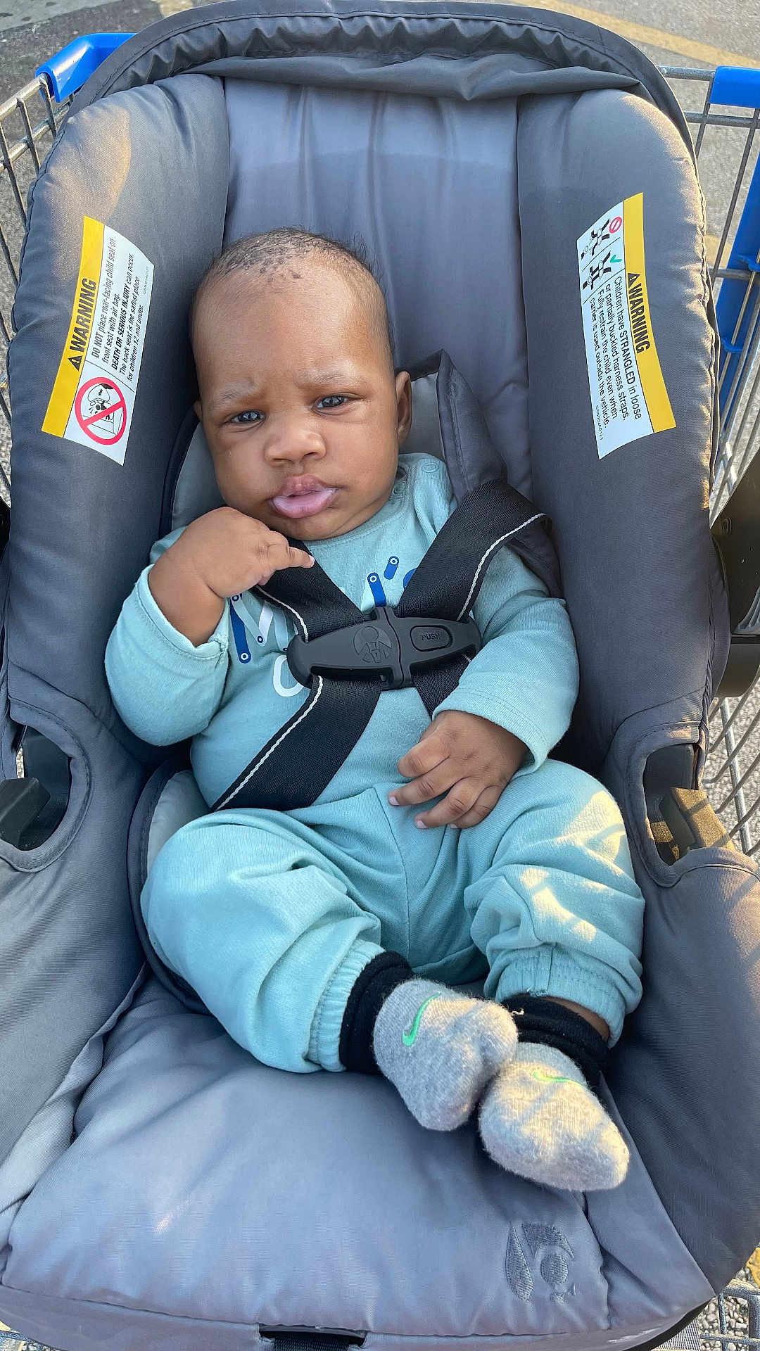 Josiah is registered to the contest to win money with this photo: baby, infant, car_seat, shopping_cart, blue_clothing, socks, seatbelt, child, cute, outdoor, straps, expression, serious, hands, feet, person, padded_seat, warning_labels, daylight, young_child