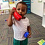 Isaiah joined the competition — help win amazing prizes! toddler, child, toy_phone, blue_toy, classroom, carpet, educational_posters, playroom, indoor, casual_clothing, shorts, white_shirt, green_shorts, young_child, learning_environment, colorful, playtime, person, floor, furniture