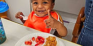 Isaiah joined the competition — help win amazing prizes! toddler, child, smiling, orange_bib, highchair, plate, food, spoon, table, indoor, happy, meal, fruit, strawberry, grain, baby, person, eating, water_bottle, clothing