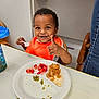 toddler, child, smiling, orange_bib, highchair, plate, food, spoon, table, indoor, happy, meal, fruit, strawberry, grain, baby, person, eating, water_bottle, clothing