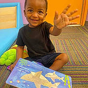 Isaiah joined the competition — help win amazing prizes! toddler, child, smiling, waving, book, reading, carpet, colorful, indoor, orange_door, black_tshirt, shorts, toy, learning, education, playful, happy, cute, person, floor