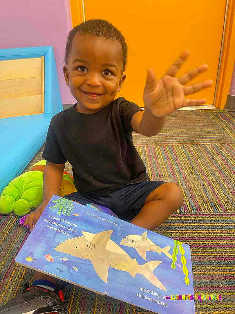 Isaiah joined the competition — help win amazing prizes! toddler, child, smiling, waving, book, reading, carpet, colorful, indoor, orange_door, black_tshirt, shorts, toy, learning, education, playful, happy, cute, person, floor