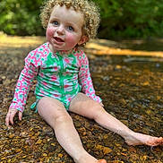 Delilah joined the competition — help win amazing prizes! toddler, child, curly_hair, blue_eyes, smiling, water, creek, rocks, outdoor, nature, sunlight, greenery, swimsuit, floral_pattern, playful, sitting, happy, summer, skin, barefoot