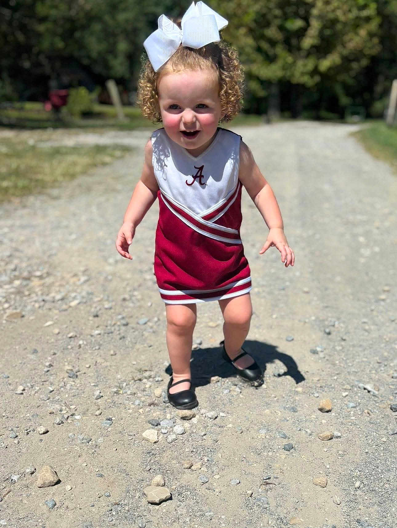 Delilah joined the competition — help win amazing prizes! toddler, child, curly_hair, white_bow, cheerleading_outfit, maroon, white, dress, shoes, gravel_path, outdoor, sunlight, shadow, happy, walking, confident, nature, trees, daytime, person