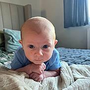 Oakley is registered to the contest to win money with this photo: baby, infant, blue_onesie, blanket, bed, pillow, headboard, curtains, window, indoors, face, hands, expression, child, soft_texture, cozy, natural_light, portrait, resting, cute