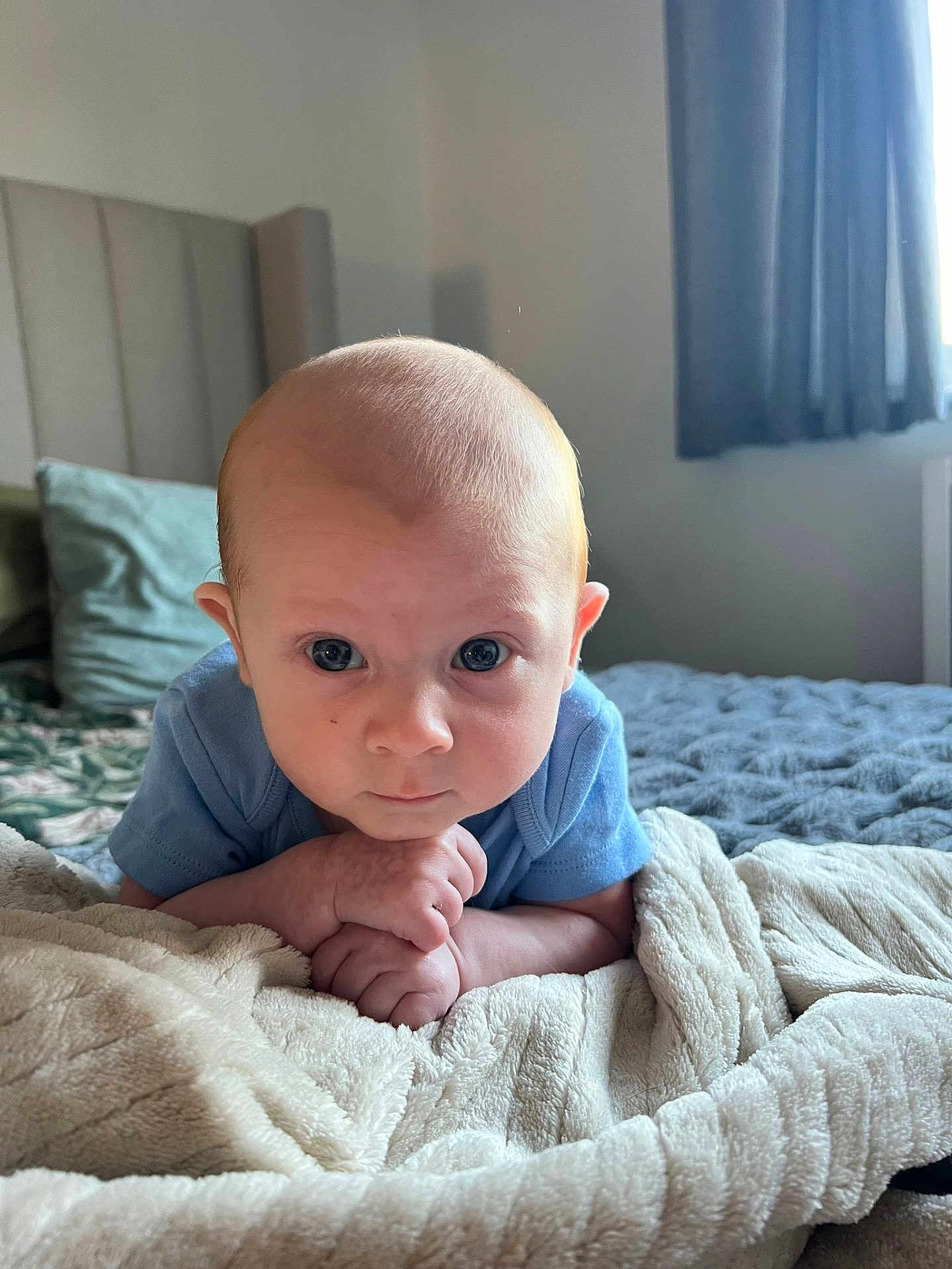 Oakley is registered to the contest to win money with this photo: baby, infant, blue_onesie, blanket, bed, pillow, headboard, curtains, window, indoors, face, hands, expression, child, soft_texture, cozy, natural_light, portrait, resting, cute