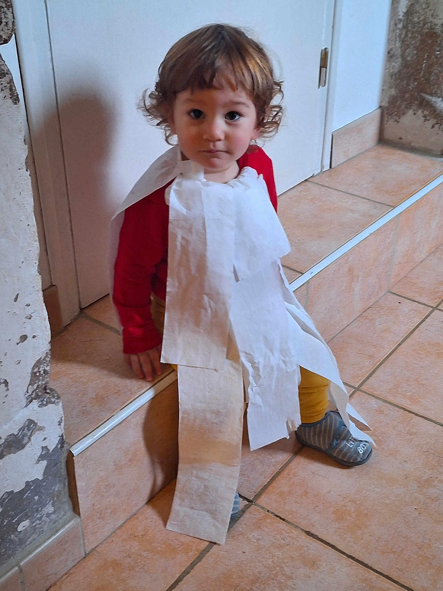 Favio a rejoint le concours — aidez-le/la à gagner de superbes lots ! toddler, child, toilet_paper, cape, red_shirt, yellow_pants, indoor, tiles, steps, curly_hair, footwear, blue_slippers, wall, door, costume, playful, portrait, sitting, expression, home
