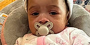 Nevaeh is registered to the contest to win money with this photo: baby, pacifier, mittens, hat, infant, cushion, seat, pink, white, clothing, face, indoors, wooden_cabinet, floral_pattern, comfort, resting, child, cute, portrait, newborn