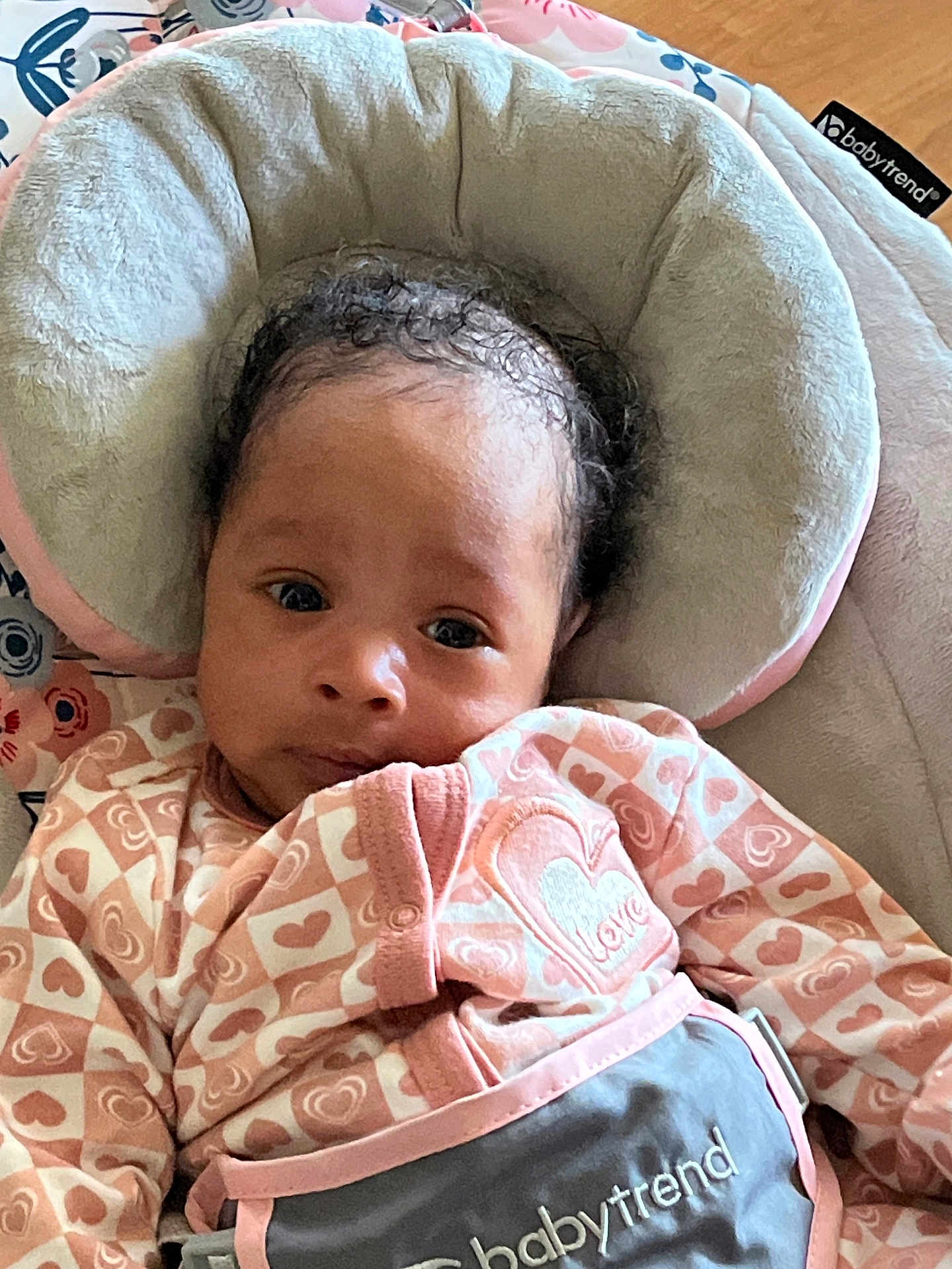 Keilani is registered to the contest to win money with this photo: baby, infant, child, curly_hair, pink_clothing, heart_pattern, love, baby_seat, plush_cushion, indoor, portrait, cute, cozy, soft_texture, wide_eyes, looking_at_camera, wooden_floor, comfortable, person, close_up