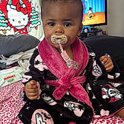 Kemani is registered to the contest to win money with this photo: baby, bed, bedroom, black, child, face, fluffy, hello_kitty, home, indoor, infant, pacifier, patterned_blanket, person, pink, portrait, robe, sitting, smiling, television