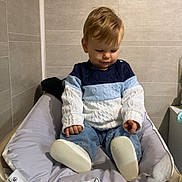 Tiago participe au concours pour gagner de l'argent avec cette photo : toddler, child, sweater, blue, white, changing_table, cushion, indoor, baby_supplies, diapers, floor, wall, tile, clothing, person, footwear, seat, baby, hair, expression