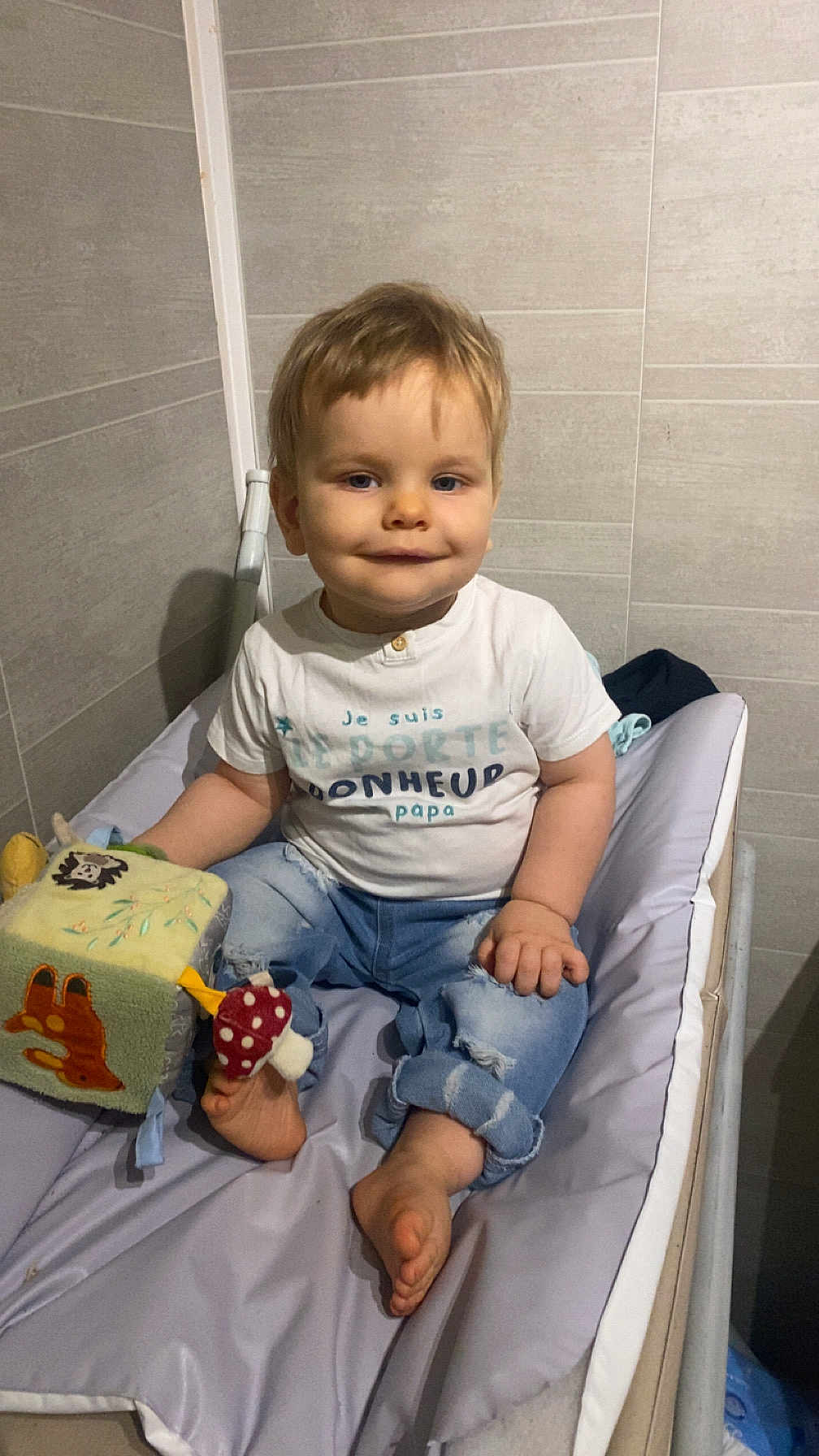 Tiago participe au concours pour gagner de l'argent avec cette photo : toddler, child, smile, toy, soft_toy, cube, white_tshirt, jeans, barefoot, changing_pad, indoor, wall, tile, baby, person, cute, sitting, play, hand, foot